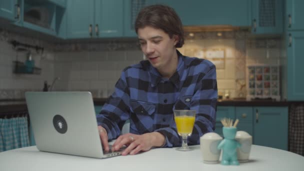 Jonge tevreden twintiger jaren mannelijke student die werkt op laptop thuis keuken. Man is blij om het werk online af te maken, zitten aan de eettafel. — Stockvideo