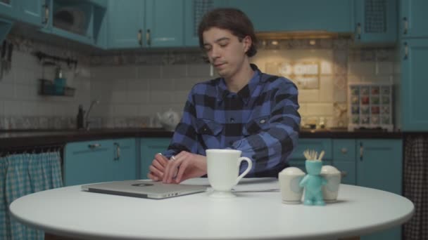 Hombre joven de 20 años con taza de té que viene y se sienta en la mesa con el ordenador portátil en la cocina azul. Estudiante masculino con documentos en papel en casa . — Vídeos de Stock