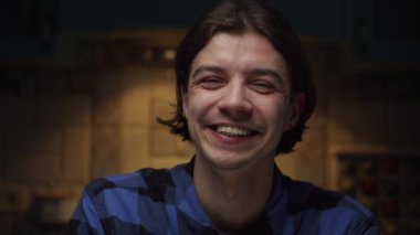 Karanlık odada kameraya yavaş çekimde bakıp gülen 20 'li yaşlardaki genç erkek portresi. Erkek yüzünde iyi bir ruh hali.. 