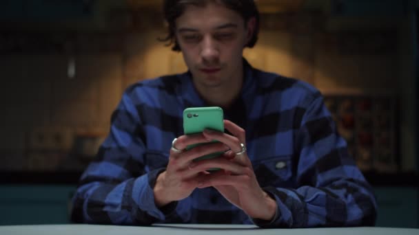 Hombre joven de 20 años usando el teléfono móvil en manos sentadas en la oscuridad. Manos de hombre con el teléfono celular en foco . — Vídeo de stock