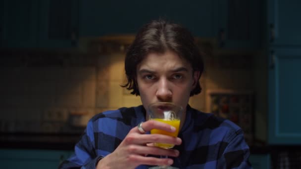 Jeune homme de 20 ans buvant du jus d'orange regardant une caméra assise dans une pièce sombre. Homme en chemise bleue boissons verre de boisson aux fruits . — Video