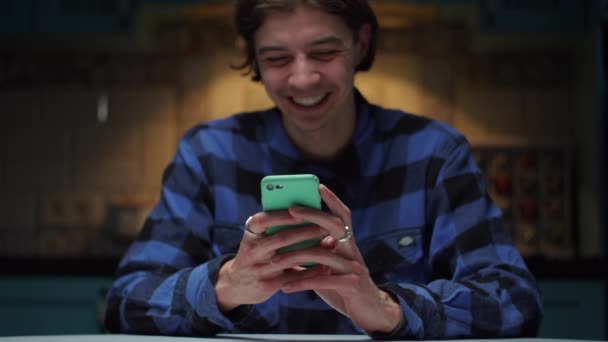 Junger lachender 20er-Jahre-Mann mit Handy in der Hand im Dunkeln sitzend. Mann lächelt und schaut auf Handy-Bildschirm. — Stockvideo