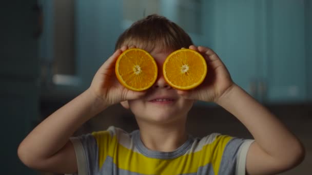 Förskolekillen håller två halvor apelsinfrukt i händerna, tar den till ögonen och ler mot kameran. Orange frukt baby ögon i slow motion. — Stockvideo