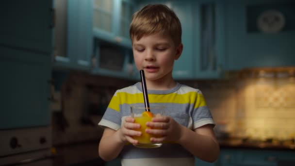 Мальчик держит в руках стакан апельсинового сока, пьет его с непластиковой соломой в замедленной съемке. Ребенок наслаждается свежим апельсиновым напитком на кухне дома . — стоковое видео