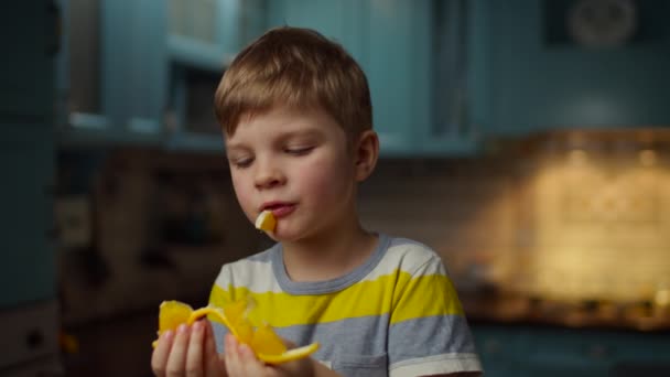Close up de menino segurando pedaço de fruta laranja nas mãos, comendo-o em câmera lenta. Kid desfrutando de laranja fresca na cozinha em casa . — Vídeo de Stock