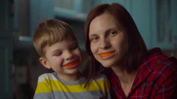 Grappige familie die thuis met sinaasappelschillen in hun mond speelt. Lachende vrouw en kleuter met oranje fruit in de mond. Oranje glimlach. — Stockvideo