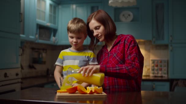 Vrouw giet sinaasappelsap in glas voor jongen. Jonge dertiger jaren moeder en zoon genieten van familietijd in de keuken. Jongen die vruchtendrank drinkt met niet-plastic stro. — Stockvideo