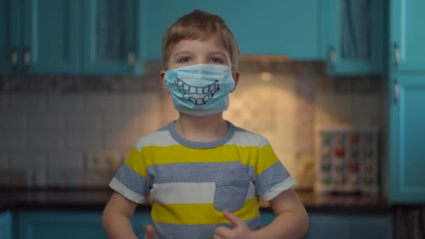 Portret van een jongen met een medisch masker met een glimlach die naar de camera kijkt en duimen laat zien. Een kind met een grappig medisch masker thuis, geïsoleerd van het virus. Stop epidemie. — Stockvideo