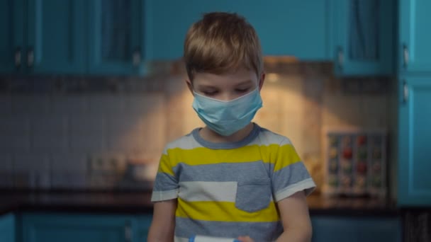 Ξανθό αγόρι με ιατρική μάσκα στο πρόσωπο μετρά τη θερμοκρασία με υπέρυθρο θερμόμετρο κοιτάζοντας την κάμερα. Παιδί με ιατρική μάσκα στο σπίτι σε απομόνωση από τον ιό. Σταμάτημα επιδημίας. — Αρχείο Βίντεο