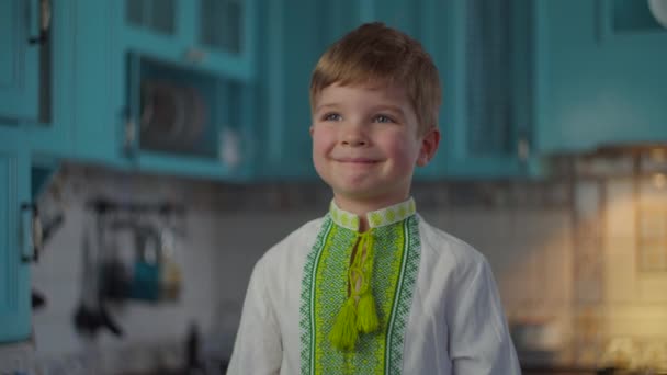 Blond pojke i broderade nationella kläder ler och visar känslor hemma på blått kök. Ung i vit stickad skjorta med etniska mönster i slow motion. — Stockvideo