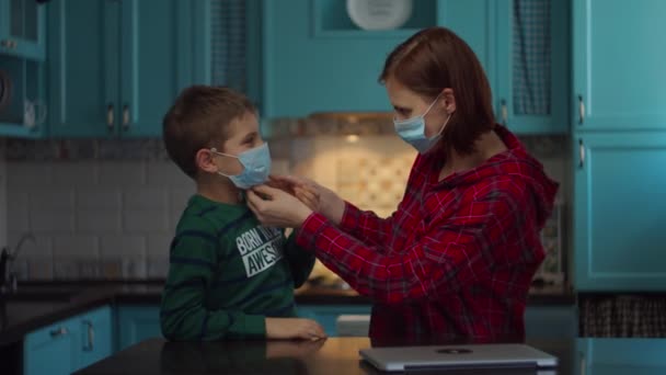 La joven madre de 30 años en máscara médica ayuda a los niños a usar máscara médica en la cara de los niños en casa. Familia con máscaras médicas en casa en aislamiento del virus . — Vídeos de Stock