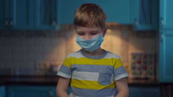 Szőke fiú orvosi maszkban méri a hőmérsékletet infravörös hőmérővel a kamerába nézve. Egy srác orvosi maszkban otthon, elszigetelve a vírustól. Járvány megállítása. — Stock videók