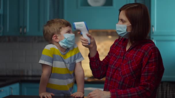 A mãe de 30 anos em máscara médica mede a temperatura de seu filho doente com termômetro infravermelho. Família em casa em auto-isolamento do vírus. Parem a epidemia . — Vídeo de Stock