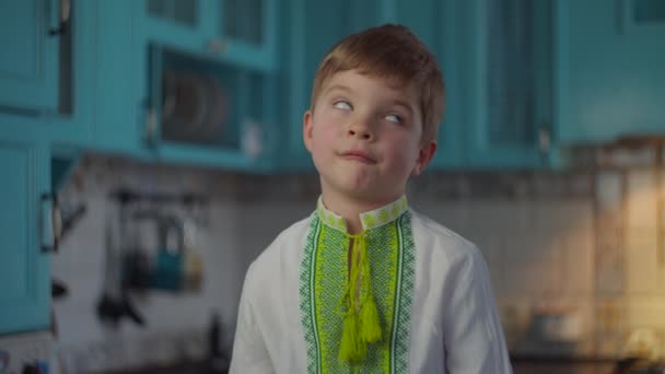 Niño rubio en ropa nacional bordada sonriendo y mostrando emociones en casa en la cocina azul. Niño en camisa de punto blanco con patrones étnicos en cámara lenta . — Vídeos de Stock