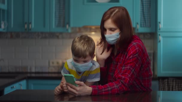 30s mother and boy in medical masks talking online by mobile phone with relatives, greeting waving hands. Family at home in self isolation from virus. Stop epidemic. — Stock Video