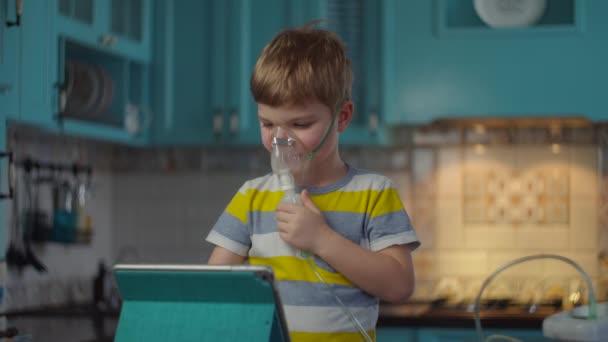 Jonge jongen die inhalator gebruikt voor de behandeling van ziekten zoals griep of respiratoire virussen en thuis naar tabletcomputer kijkt. Kind behandeld hoesten met medicijn inhalator masker op keuken.. — Stockvideo