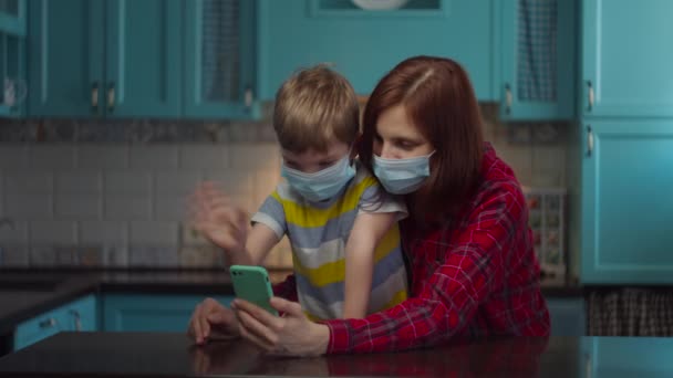 親戚と携帯電話でオンラインで話す医療マスクの30代の母親と男の子、手を振って挨拶。家庭でのウイルスからの自己分離。流行を止め. — ストック動画