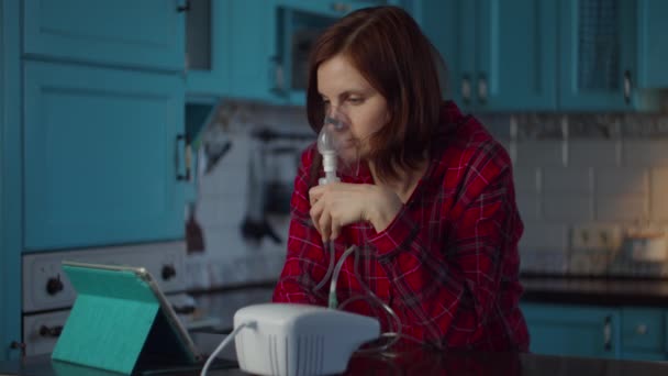 Mujer joven de 30 años con camisa roja que usa inhalador para el tratamiento de enfermedades como la gripe o los virus respiratorios y observa la computadora de la tableta en casa. Mujer que trata la tos con la máquina inhaladora de medicamentos . — Vídeo de stock