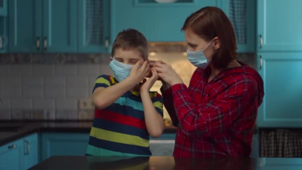 Mladé třicátnice matka v lékařské masce pomáhá dítě nosit lékařskou masku na dětském obličeji doma. Žena a chlapec v maskách, dívající se do kamery. Rodina v lékařských maskách doma v izolaci od viru. — Stock video