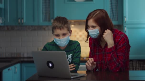 In de 30 's moeder en jongen met medische maskers kijkend naar laptop scherm en online pratend met familieleden, zwaaiende handen begroetend. Familie met medische maskers thuis, geïsoleerd van het virus. Stop epidemie. — Stockvideo