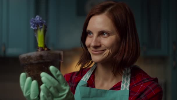 Νεαρή 30άρα γυναίκα με ποδιά και γάντια θαυμάζοντας το λουλούδι σε διάφανη κατσαρόλα στα χέρια σε αργή κίνηση. Lady απολαμβάνει εγχώρια διαδικασία φύτευσης στην κουζίνα — Αρχείο Βίντεο