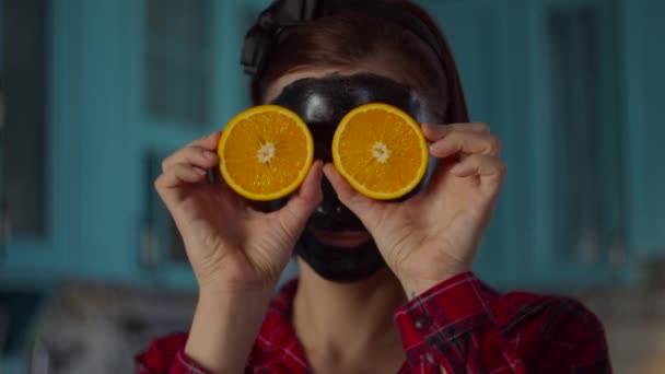 Smiling 30s woman with black cosmetic mask on her face holding orange fruit halves near eyes looking at camera. Woman beauty routine on blue kitchen. Close up. — Stock Video