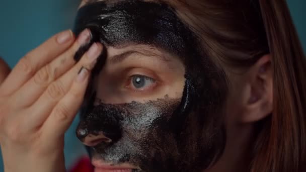 Mujer joven de 30 años con aro negro en la cabeza aplicando máscara cosmética negra en su cara. Mujer rutina de belleza en casa en la cocina azul. De cerca. . — Vídeo de stock