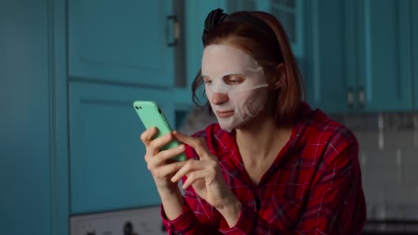 Junge Frau in den 30er Jahren mit weißer Kosmetikmaske auf dem Gesicht, zu Hause mit dem Handy. Frau schönheit routine auf blau küche. — Stockvideo