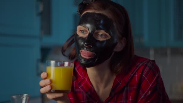 Jonge vrouw uit de 30 met een zwart cosmetisch masker op haar gezicht... die een glas sinaasappelsap drinkt terwijl ze naar de camera kijkt. vrouw schoonheid routine op blauw keuken. — Stockvideo