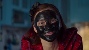 Yüzünde siyah kozmetik maske olan komik 30 'lu yaşlarda bir kadın kameraya bakarak yüzünü buruşturuyor. Mavi mutfakta maske takan komik suratlı bir kadın. Kapat.