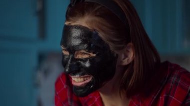 Yüzünde siyah kozmetik maske olan 30 'lu yaşlardaki komik bir kadın evde yüzünü buruşturuyor. Mavi mutfakta maske takan komik suratlı bir kadın. Kapat.