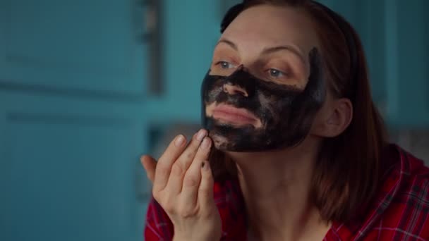 頭の上に黒いフープを持つ若い30代の女性は、彼女の顔に黒い化粧品マスクを適用します。青いキッチンで自宅で女性の美しさルーチン。中発. — ストック動画