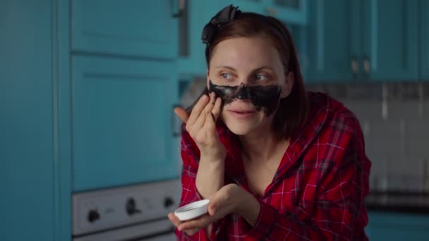 Молода 30-ті жінка з чорним кільцем на голові наносить чорну косметичну маску на обличчя. Жіноча рутина краси вдома на блакитній кухні . — стокове відео