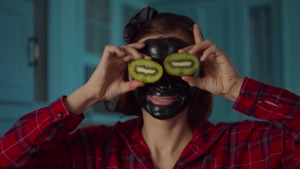 Mujer de 30 años sonriente con máscara cosmética negra en su cara sosteniendo mitades de kiwi cerca de los ojos mirando a la cámara. Mujer rutina de belleza en cocina azul . — Vídeos de Stock