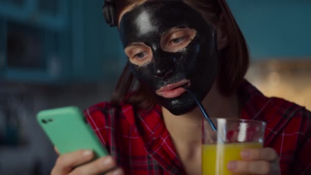 A 30-as éveiben járó fiatal nő fekete kozmetikai maszkkal az arcán narancslevet iszik nem műanyag szívószállal és mobiltelefont használ. Nő szépség rutin kék konyha. — Stock videók