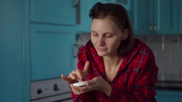 Jonge vrouw uit de 30 met een zwarte hoepel op haar hoofd, die een zwart cosmetisch masker op haar gezicht aanbrengt. vrouw schoonheid routine thuis op blauw keuken. — Stockvideo