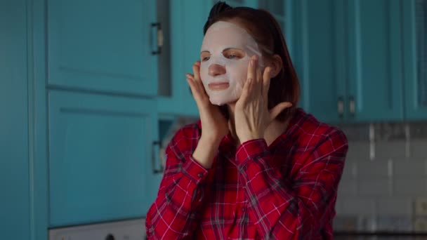 Jeune femme des années 30 avec un cerceau noir sur la tête appliquant un masque cosmétique blanc sur son visage. Femme beauté routine à la maison sur cuisine bleue . — Video