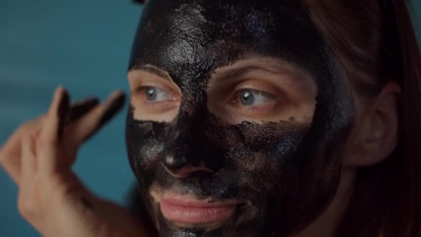 Young 30s woman with black hoop on head applying black cosmetic mask on her face. Woman beauty routine at home on blue kitchen. Close up. — Stock Video