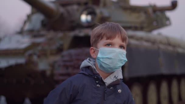 Αγόρι με ιατρική προστατευτική μάσκα στέκεται δίπλα σε στρατιωτική μηχανή κατά τη διάρκεια πανδημίας ξέσπασμα του coronavirus COVID-19 και φοβάται κοιτάζει γύρω του. Επείγουσα κατάσταση καραντίνας και στρατιωτικός νόμος για την καταπολέμηση του κορωναϊού. — Αρχείο Βίντεο