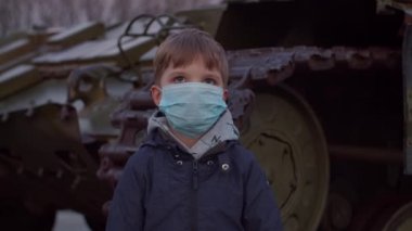 Tıbbi koruyucu maskeli çocuk Coronavirus COVID 19 salgını sırasında askeri makinenin yanında duruyor ve etrafı izliyor. Koronavirüsle savaşmak için acil karantina ve sıkıyönetim. Kapat.