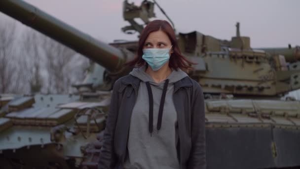 Молодая женщина в медицинской защитной маске стоит у военной машины во время пандемической вспышки коронавируса COVID-19 и испуганно смотрит вокруг. Карантинная чрезвычайная ситуация и военное положение для борьбы с коронавирусом . — стоковое видео