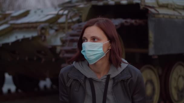 Женщина в медицинской защитной маске стоит у военной машины во время пандемической вспышки коронавируса COVID-19 и испуганно оглядывается вокруг. Карантин и военное положение для борьбы с коронавирусом. Закрыть — стоковое видео