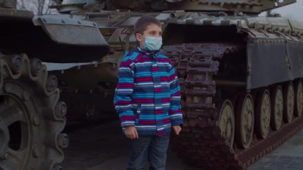 Мальчик в медицинской защитной маске стоит у военной машины во время пандемической вспышки коронавируса COVID-19 и наблюдает вокруг. Карантин национальной чрезвычайной ситуации и военного положения для борьбы с коронавирусом . — стоковое видео