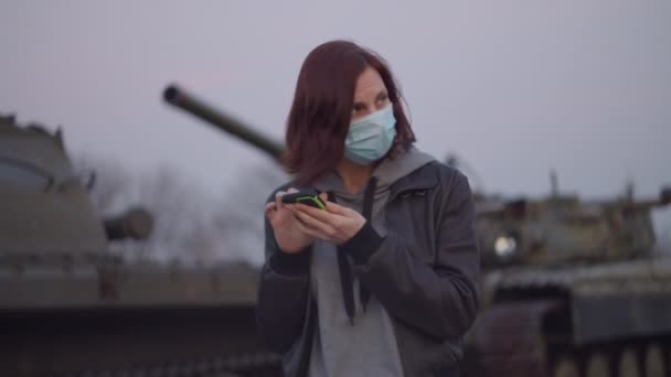 Young woman in medical protective mask stands by military machine during pandemic outbreak of coronavirus COVID-19 and scared looks around. Quarantine emergency and martial law to combat coronavirus. — Stock Video