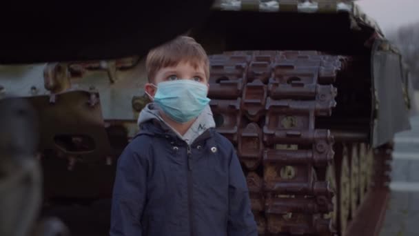 Menino em máscara protetora médica fica ao lado de máquina militar durante surto pandêmico de coronavírus COVID-19 e observando ao redor. Quarentena lei nacional de emergência e marcial para combater o coronavírus . — Vídeo de Stock