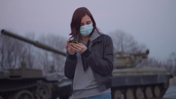 Jonge vrouw met medisch beschermend masker staat bij de militaire machine tijdens een pandemische uitbraak van het coronavirus COVID-19 en bang kijkt om zich heen. Quarantaine nood- en krijgswet ter bestrijding van het coronavirus. — Stockvideo