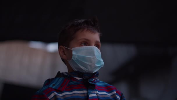 Мальчик в медицинской защитной маске стоит в укрытии во время пандемической вспышки коронавируса COVID-19 и наблюдает вокруг. Карантин национальной чрезвычайной ситуации и военного положения для борьбы с коронавирусом. Закрыть — стоковое видео