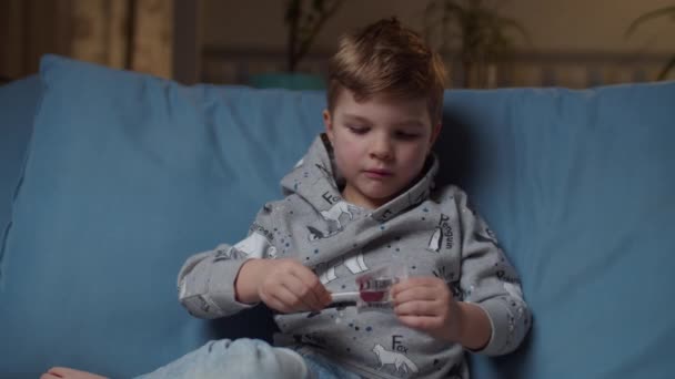 Young boy opening sweets and take lollipop to the mouth sitting on cozy couch at evening night. Kid enjoying hard candy and smiling looking at camera. — Stock Video