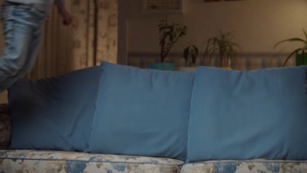 Kind in Jeans springt auf Couch im Wohnzimmer. Kind macht akrobatische Bewegungen auf Couch. Blonder Junge albert abends auf gemütlichem Sofa herum. — Stockvideo