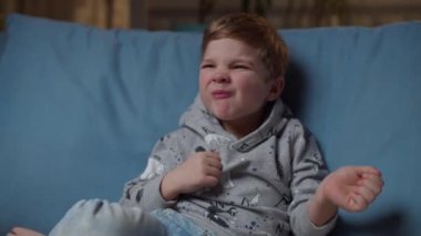 Sarışın çocuk akşam odasındaki rahat koltukta oturuyor. Kotlu çocuk oturma odasında tek başına oynuyor. Komik çocuk kanepede dans ediyor.. 